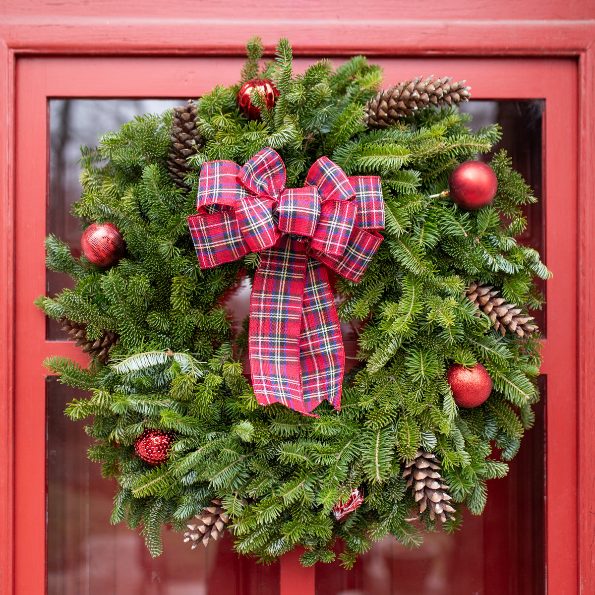 A festive evergreen wreath with pinecones and red ornaments hanging on a red front door. The wreath features a large red and green plaid bow in the center.